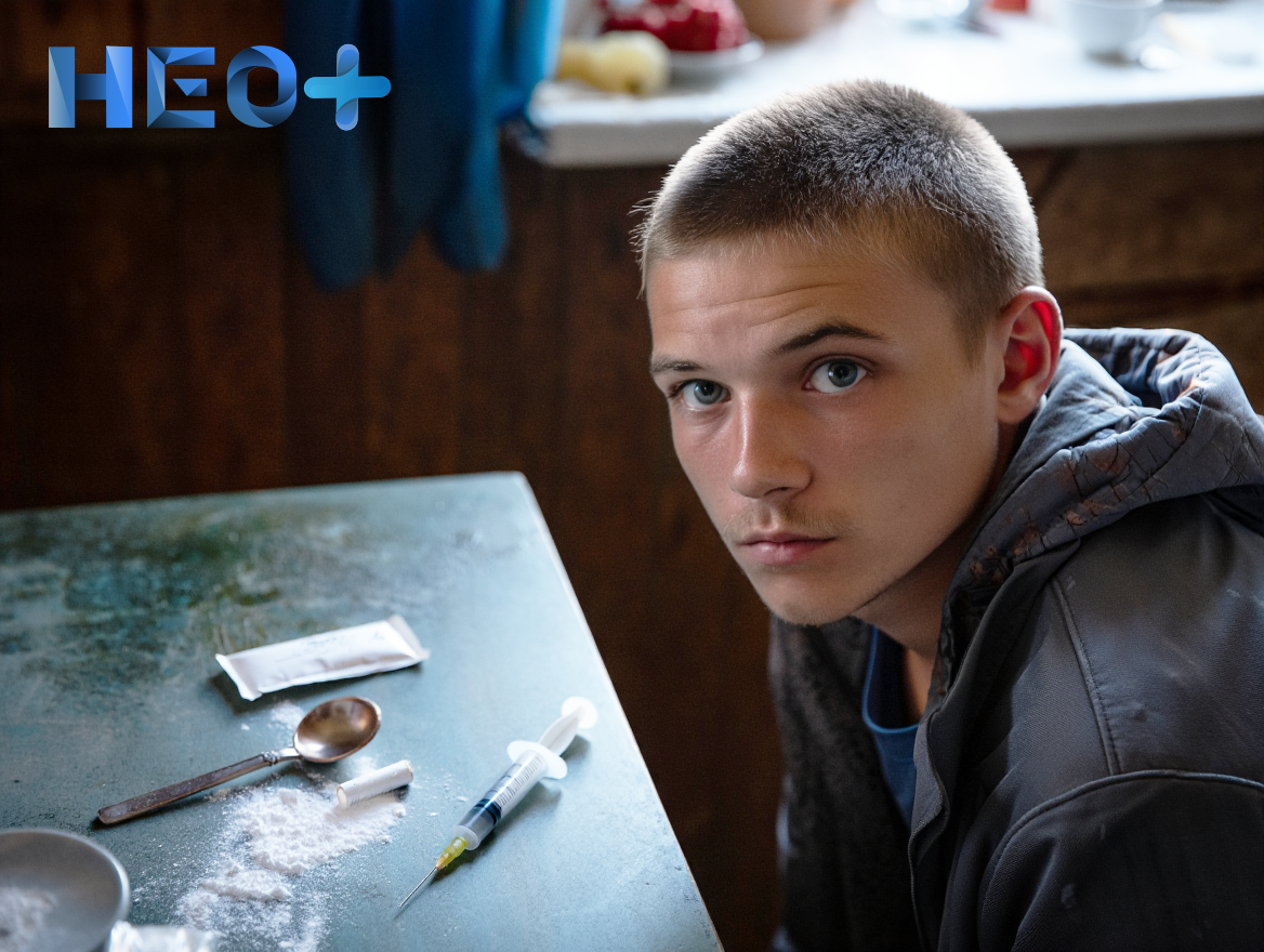 Молодой парень сидит за столом на котором шприц, ложка и порошок